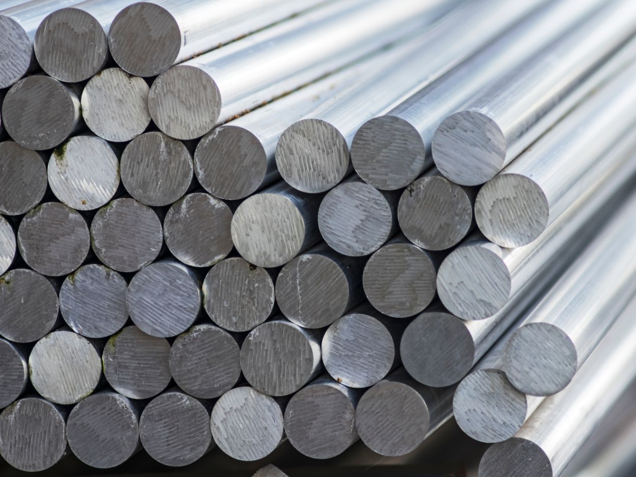 Barras cilíndricas de metal empilhadas em estoque industrial, representando diferentes tipos de aço utilizados na fabricação.