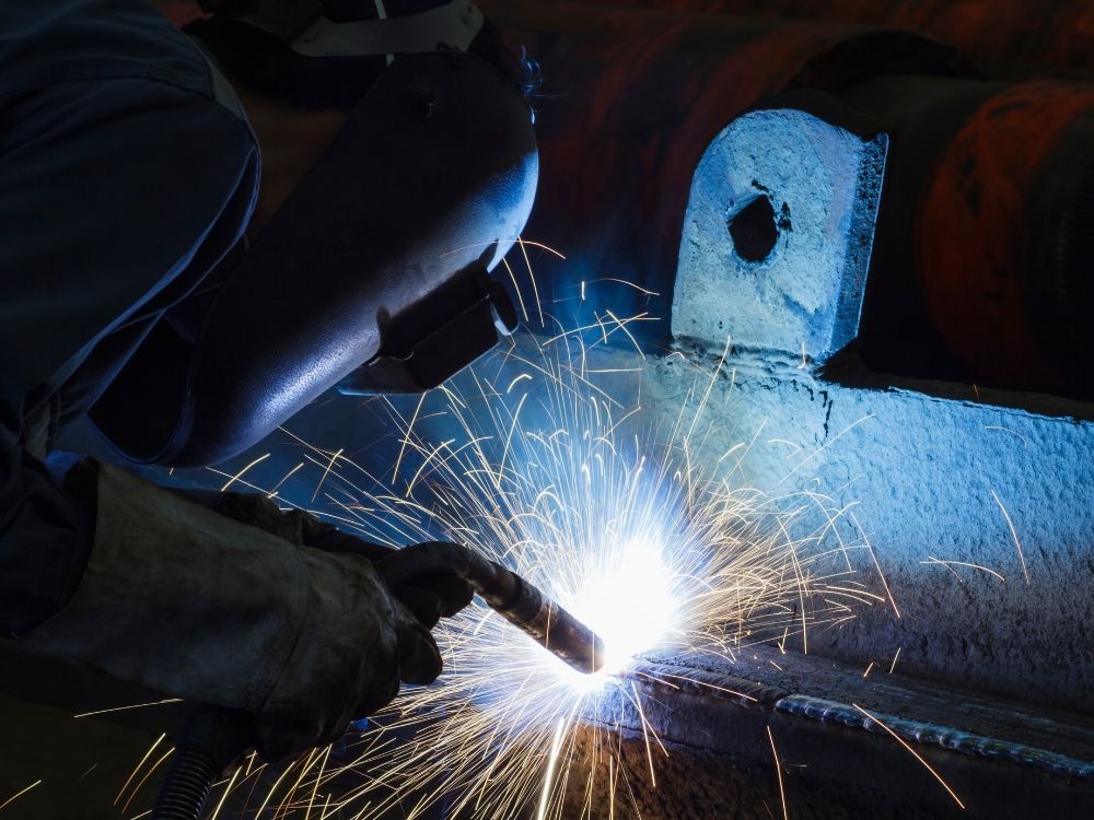 Soldador usando máscara de proteção enquanto realiza soldagem MIG em peça industrial, exemplificando cenários reais que ajudam a entender se Solda MIG é boa.