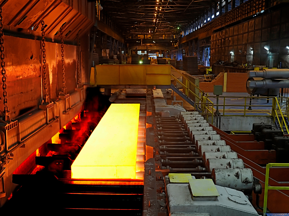Placa metálica aquecida passando por equipamentos industriais, representando uma etapa do processamento do aço.