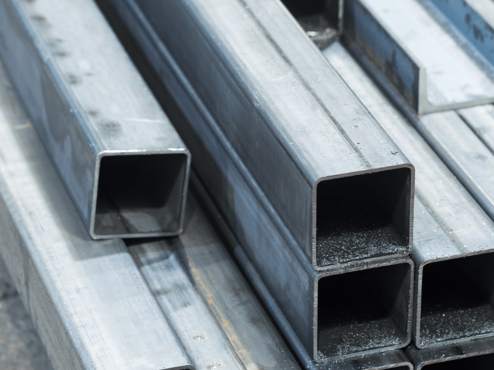 Tubos metálicos de seção quadrada empilhados, com superfícies prateadas e extremidades abertas, usados em aplicações industriais e estruturais.