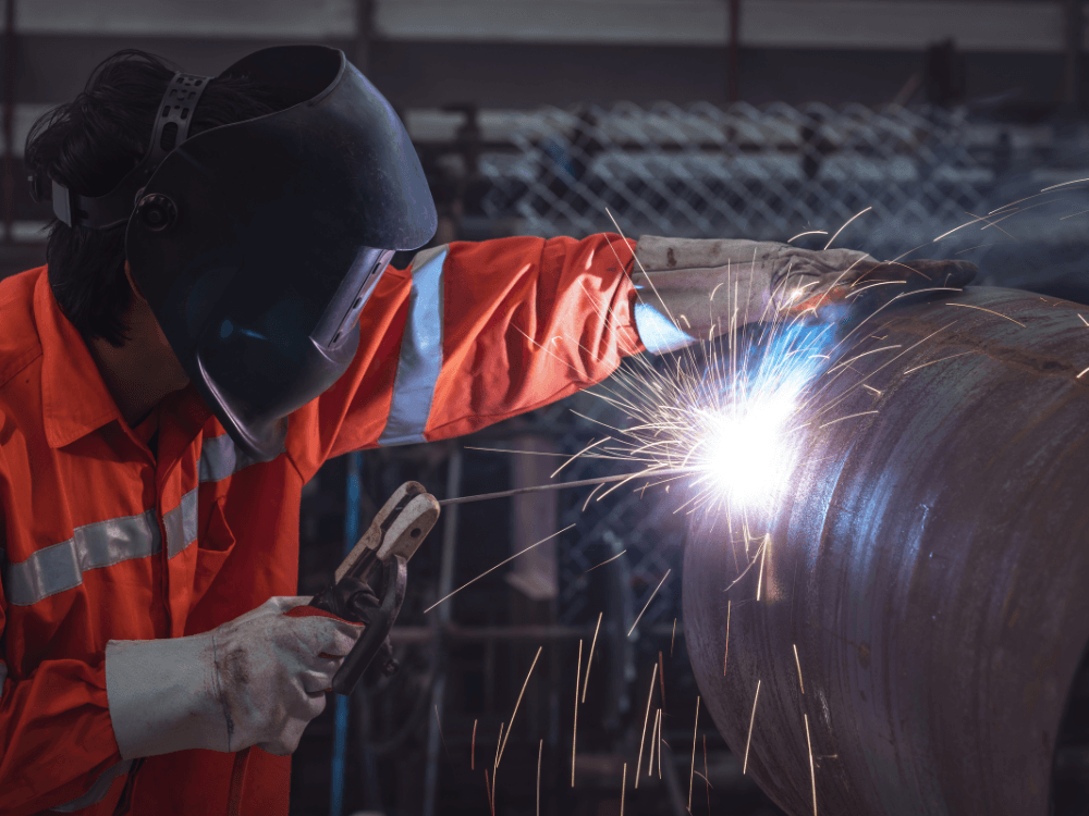 Trabalhador realiza soldagem em uma grande peça metálica cilíndrica, com máscara de proteção e faíscas durante o processo industrial. Tudo para explicar o que é ferro fundido.