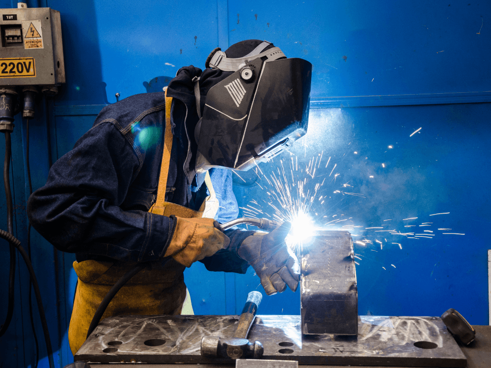 Profissional com máscara de solda e luvas trabalha em uma peça metálica sobre uma bancada, com faíscas iluminando o ambiente industrial.