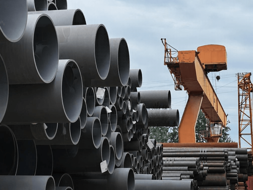 Pilhas de tubos metálicos de grande diâmetro estão organizadas em um pátio industrial próximo a estruturas de movimentação de cargas.